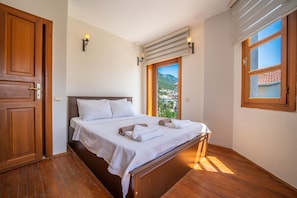 A bedroom with a wooden bed, white linens, a view of a mountain, and a window with a view of a building. - Villa with stunning sea view and private pool (Kaş)