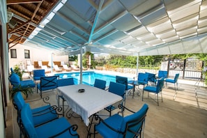 Zona de piscina exterior amb caires blaus i taula, envoltada d'un porxo cobert.