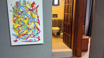 Um corredor com porta de madeira que dá para um banheiro, um quadro abstrato e colorido na parede e um piso ladrilhado.