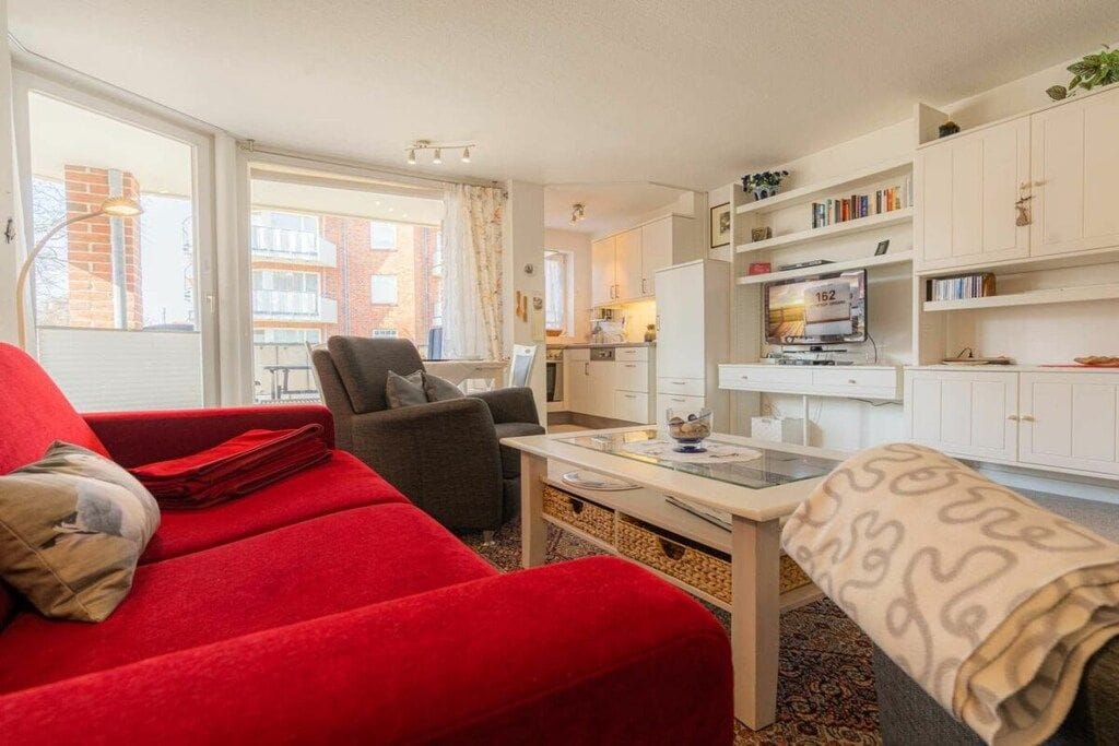 Een woonkamer met een rode bank, een grijze fauteuil, een glazen salontafel en een witte kast met boeken.