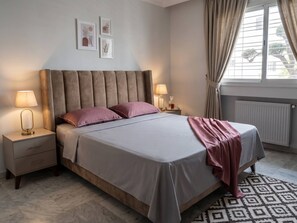 A bedroom with a bed, nightstands, lamps, a radiator, and a window with curtains. - Gardens of Carthage quiet apartment (Tunis)