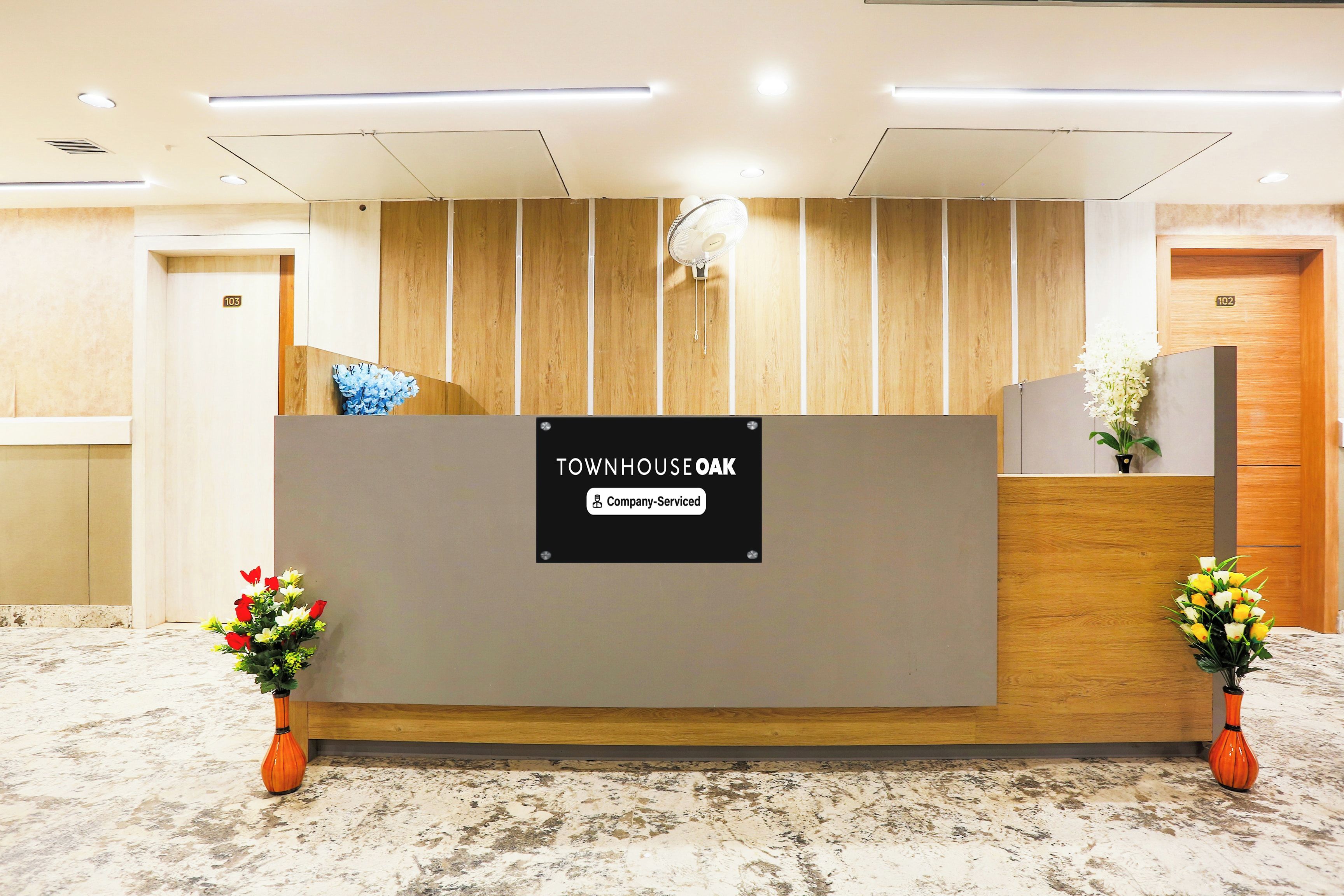 Ein Empfangsschalter mit Firmenlogo, flankiert von zwei Blumenarrangements, und eine Wand aus Holzverkleidung im Hintergrund.