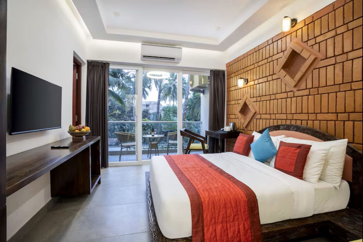 Quarto de hotel com cama, televisão, escrivaninha e varanda vista para palmeiras.