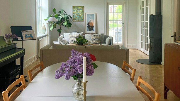 Et spiseområde med et hvidt bord, stole, en vase med lilla blomster og et klaver til venstre.