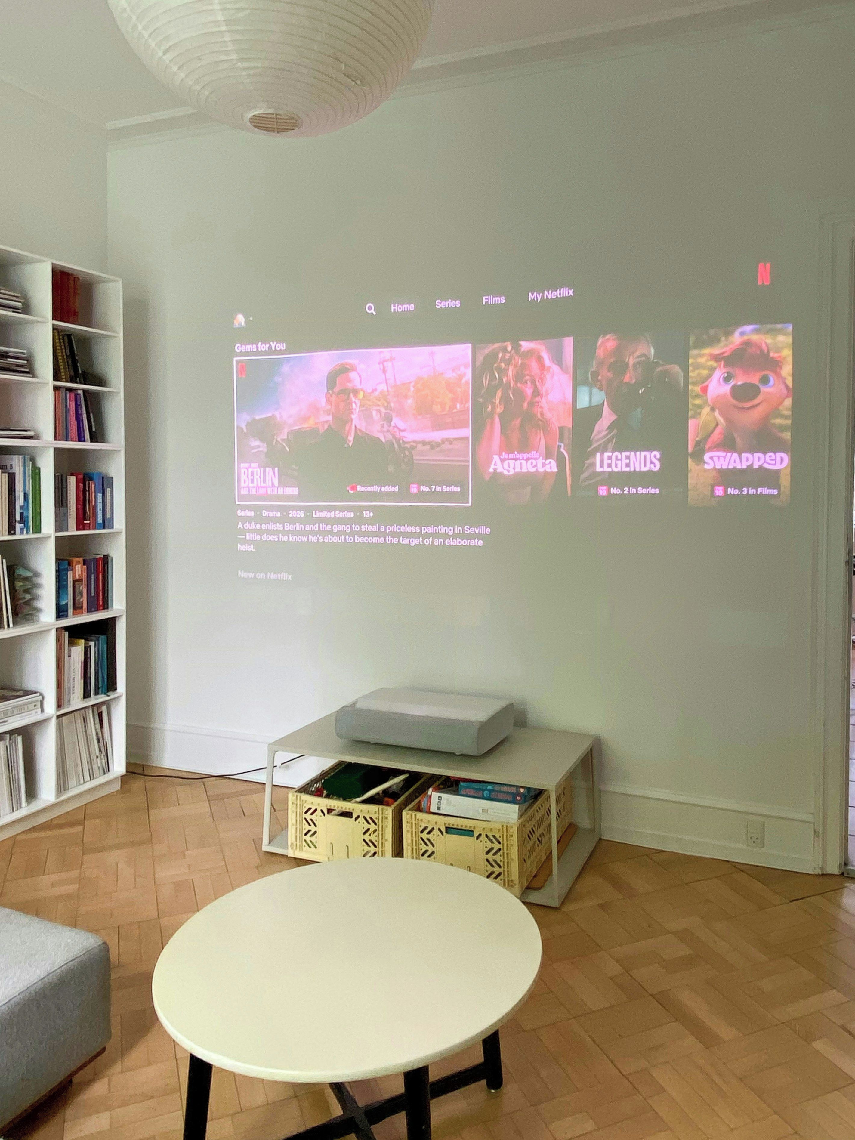 Een kamer met een witte muur, een boekenkast, een tafel en een beamer die een Netflix-interface toont.