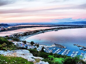 A coastal marina with boats docked, a bridge connecting to the mainland, and a hilly landscape in the background.