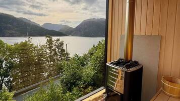 A wooden deck with a view of a lake and mountains, a wood-burning stove, and a wooden bench.