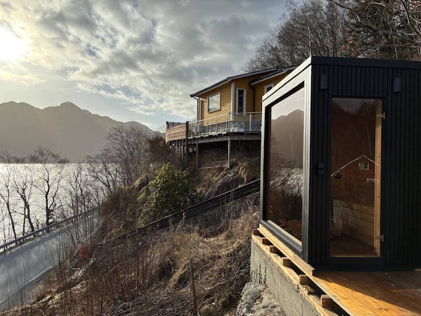 Une maison moderne dotée d’une terrasse offrant une vue sur un plan d’eau et sur des montagnes en arrière-plan.