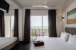 Une chambre d’hôtel avec un lit, un ventilateur de plafond, une grande fenêtre avec des rideaux, une vue sur la mer et un tableau au mur.