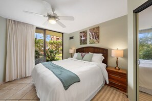 A hotel room with a bed, a ceiling fan, a nightstand with a lamp, and a view of greenery outside. - Beach Front & Just Steps to Pools & Hot Tub (Kihei)