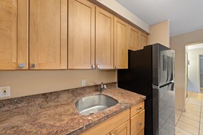 A kitchen with wooden cabinets, a granite countertop, a stainless steel sink, and a black refrigerator. - Beach Front & Just Steps to Pools & Hot Tub (Kihei)