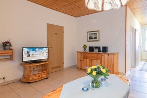 A room with a television on a wooden stand, a cabinet, a table with a vase of flowers, and a door. - 2 in the house Sturmmöwe (ID 120) (St. Peter-Ording)