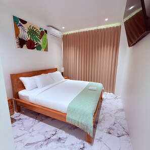 Quarto de hotel com cama de madeira, manta verde, piso com padrão marmorizado, ventoinha no teto e televisão fixada na parede.