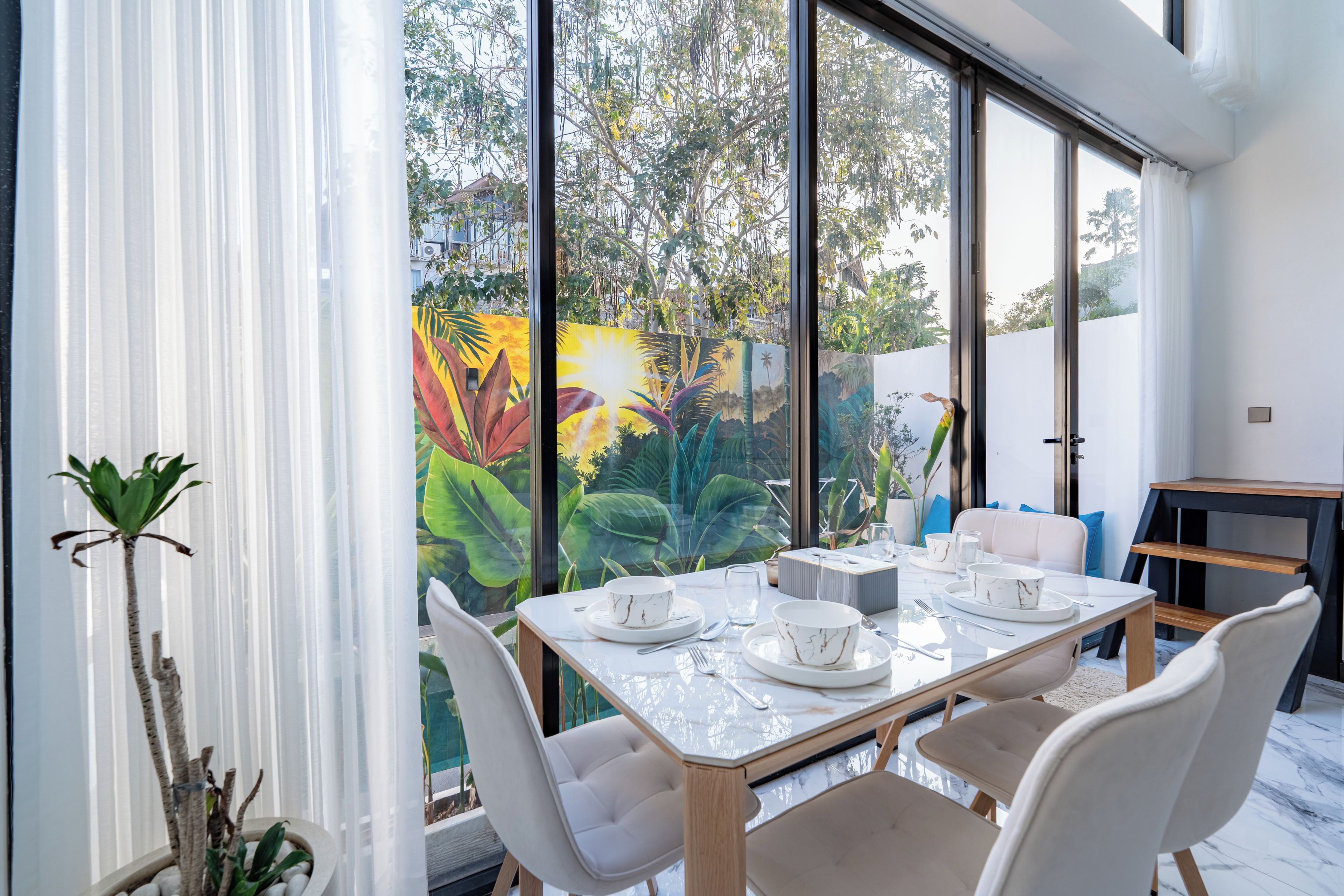 A dining area with a table set for four, large windows, and a view of greenery outside.