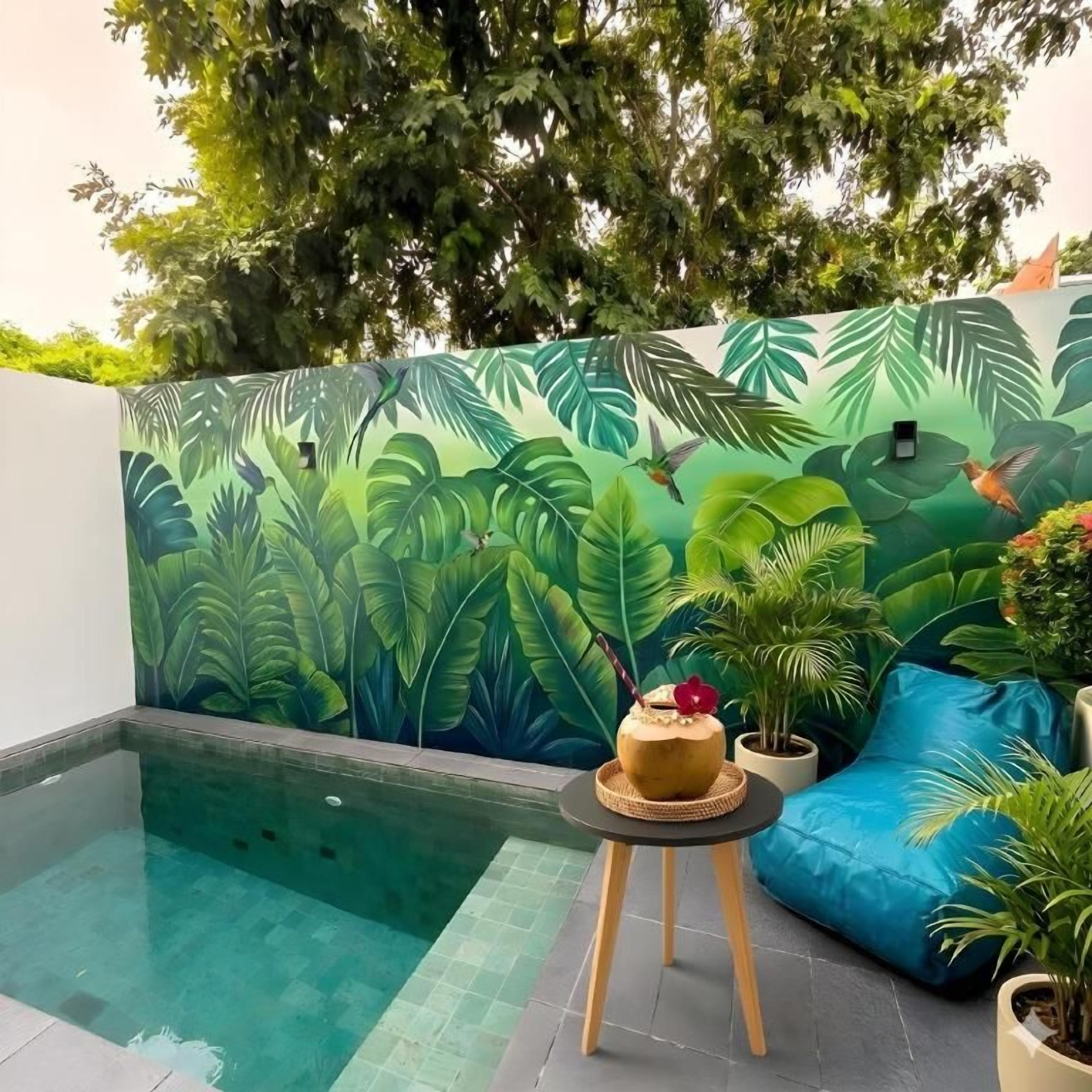 A pool area with a mural of tropical leaves and birds, a small round table with a drink, and a blue cushioned seating area.
