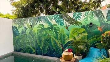 A pool area with a mural of tropical leaves and birds, a small round table with a drink, and a blue cushioned seating area.