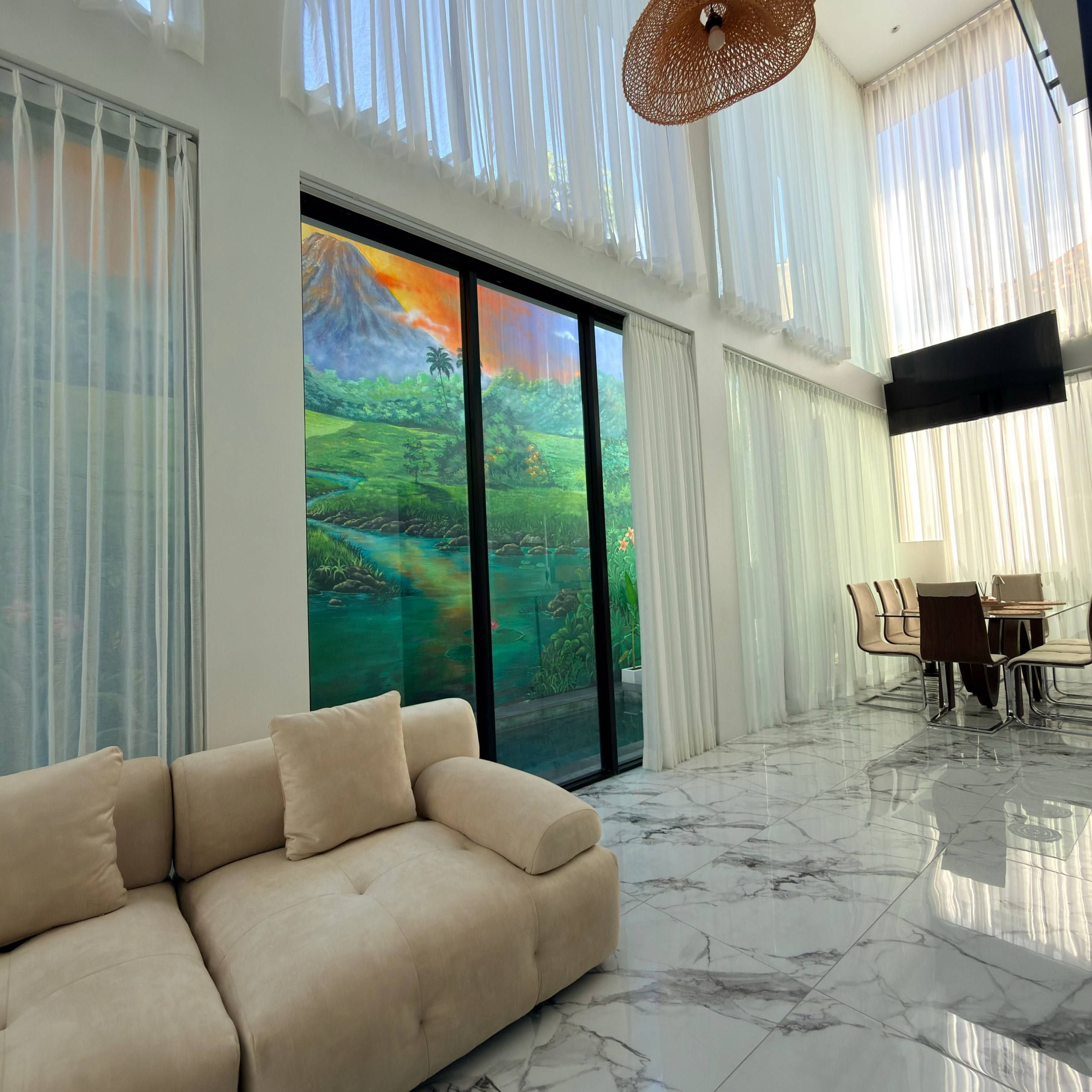 A modern living room with a beige sofa, a dining area with a table and chairs, and a large window with a view of a green landscape.