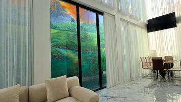A modern living room with a beige sofa, a dining area with a table and chairs, and a large window with a view of a green landscape.