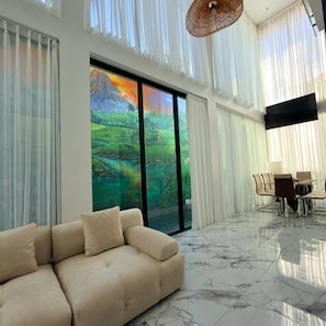 A modern living room with a beige sofa, a dining area with a table and chairs, and a large window with a view of a green landscape. - Sun On the Hill Villa by Betterplace (Nusa Dua)