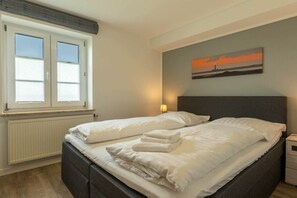 Ein Doppelbett mit weißer Bettwäsche und Kopfteil, ein Fenster mit Jalousien, eine Heizung und ein Wandbild.