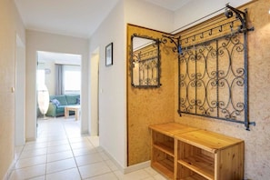Ein Flur mit einer Holzbänklchen, einem Metallgestell mit aufgehängten Gegenständen und Blick in einen Wohnbereich mit Sofa und Lampe.