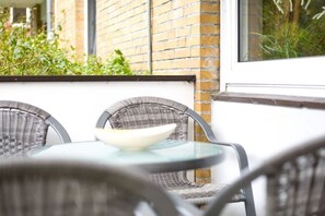 A glass-top table with two wicker chairs, a white bowl, and a brick wall with a window in the background. - 22 in the Sandkamp building (ID 051) (St. Peter-Dorf)