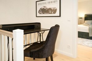 Quarto moderno com área de refeições, destacando uma cadeira escura e uma mesa, um quadro na parede e uma cama com cabeceira ao fundo.