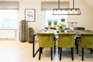 Uma sala de jantar com uma mesa posta para seis pessoas, rodeada por seis cadeiras verdes, um lustre pendurado acima e um quadro na parede.