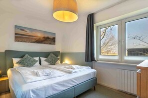 A neatly made bed with pillows, a bedside lamp, a window with curtains, and a painting on the wall. - 1 in the Westmarken building (ID 197) (St. Peter-Ording)