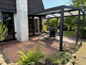 Ein überdachter Patio-Bereich mit Tisch und Stühlen, ein Kiesweg und umliegendes Grün.