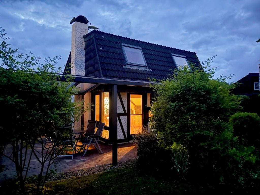 Ein Haus mit geneigtem Dach, Schornstein und einer überdachten Terrasse mit Stühlen.