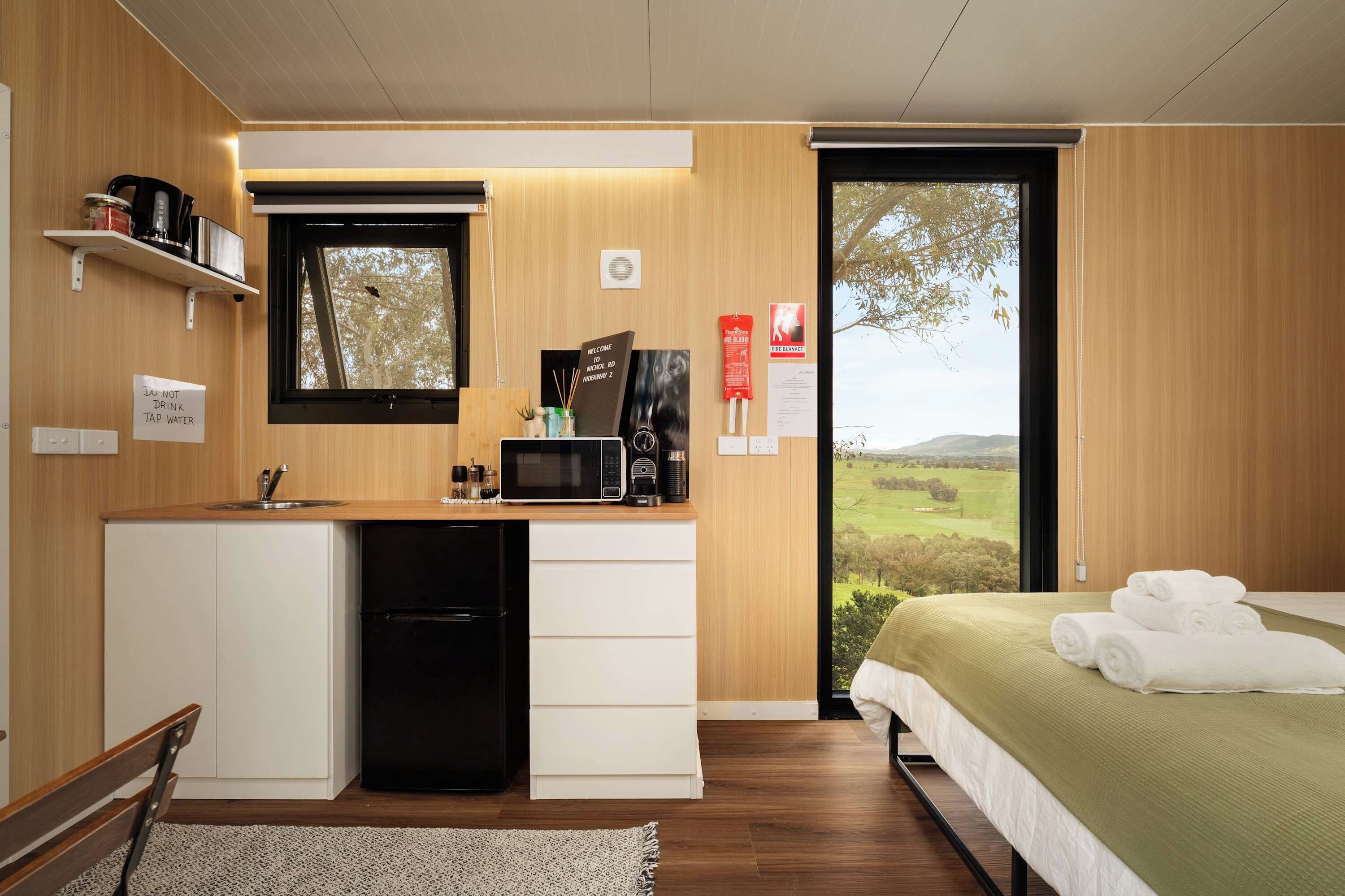 Ein kompaktes Hotelzimmer mit Küchenzeile, einem Bett mit grüner Bettdecke und Blick auf die Landschaft durch ein großes Fenster.