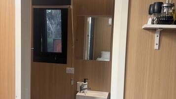 A compact bathroom with a toilet, sink, and mirror. There is a shelf with bottles and a small cabinet.