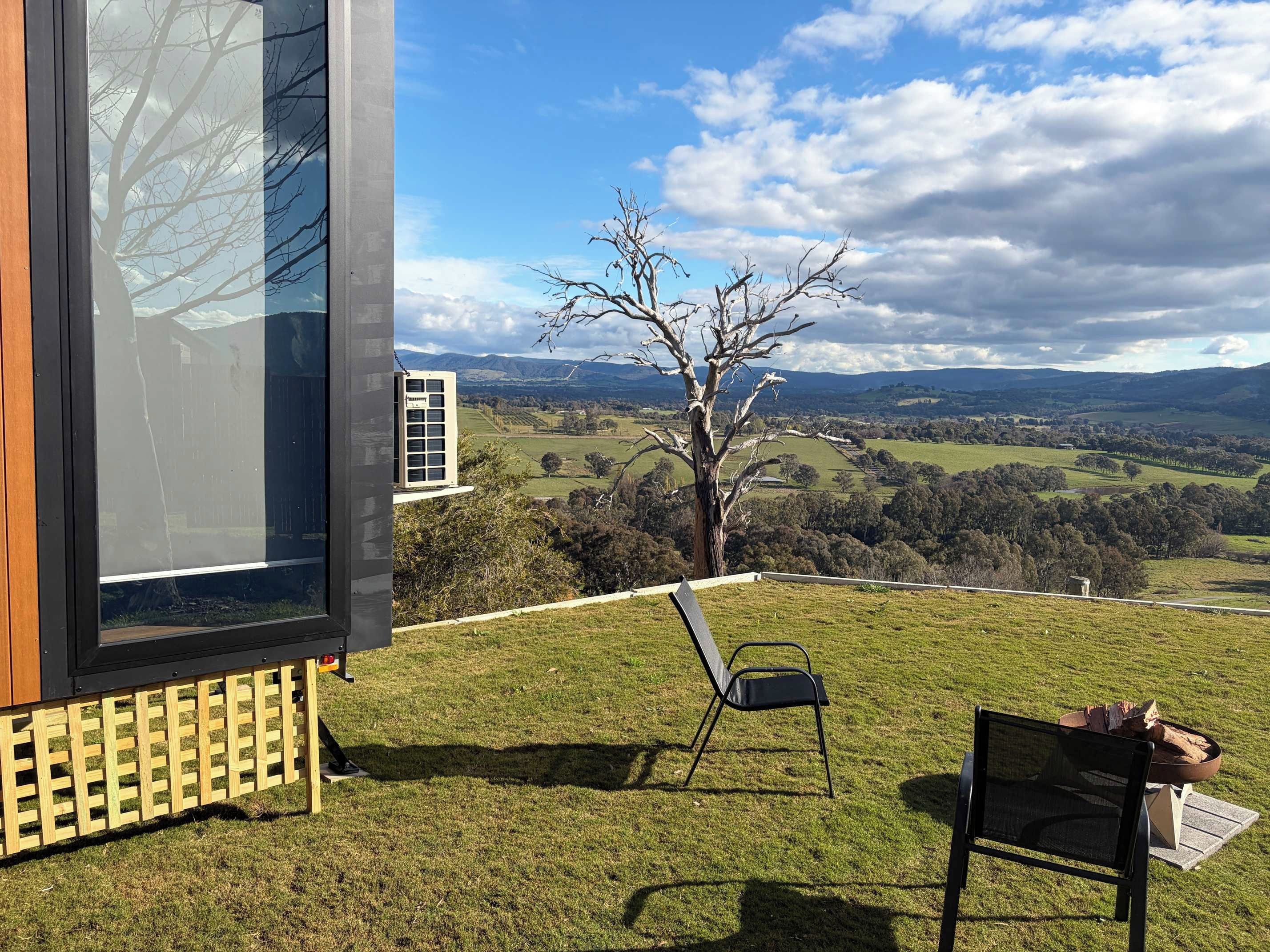 Un bâtiment moderne doté d’une grande baie vitrée, d’une terrasse en bois, d’une chaise en métal et d’un foyer.