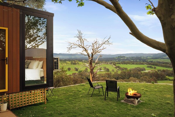 A modern glass structure with a bed and a fire pit, overlooking a green landscape.