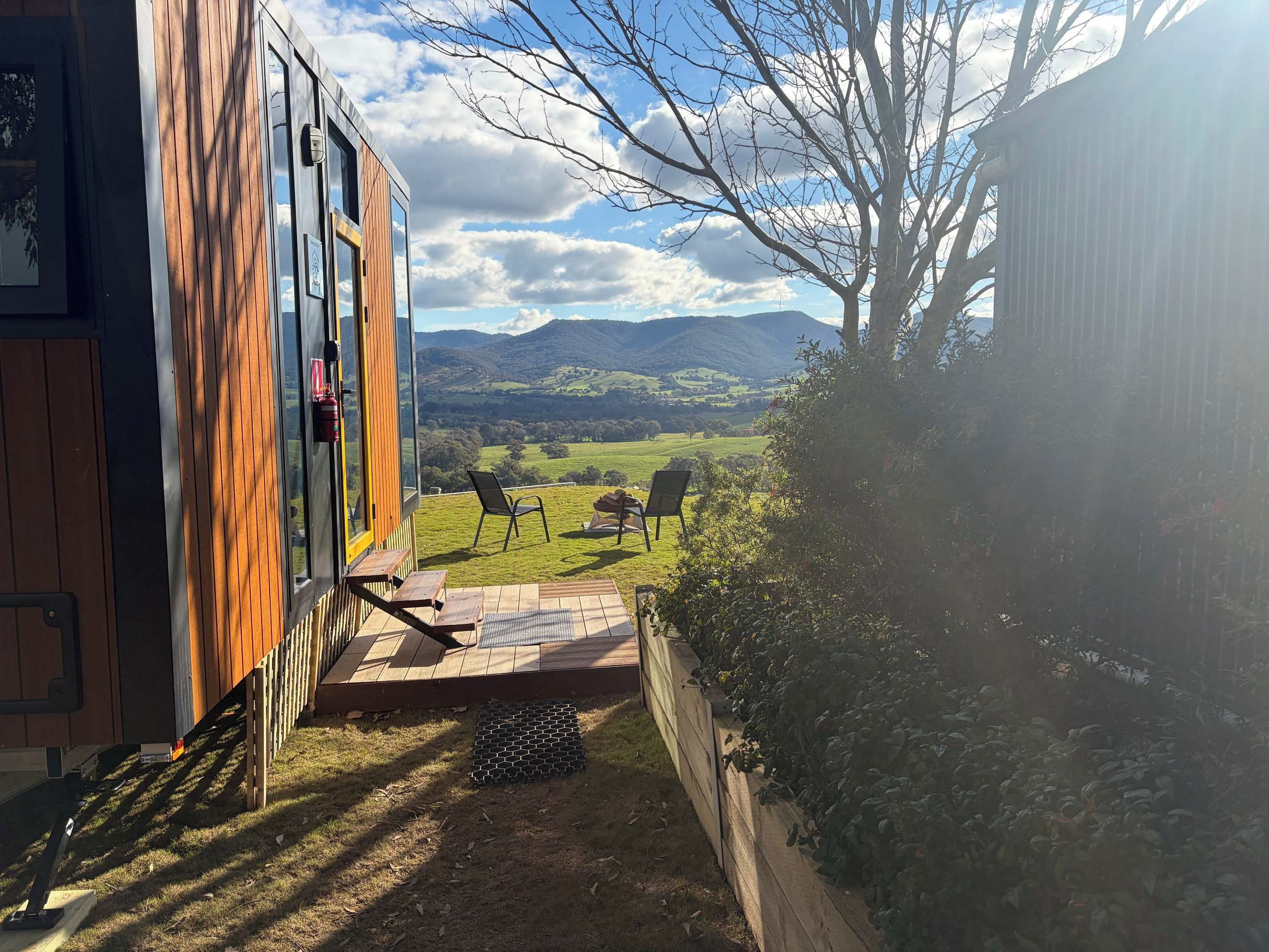Ein modernes Gebäude mit Holzfassade, einer Terrasse mit Stühlen und Blick auf eine grüne Landschaft mit Bergen im Hintergrund.