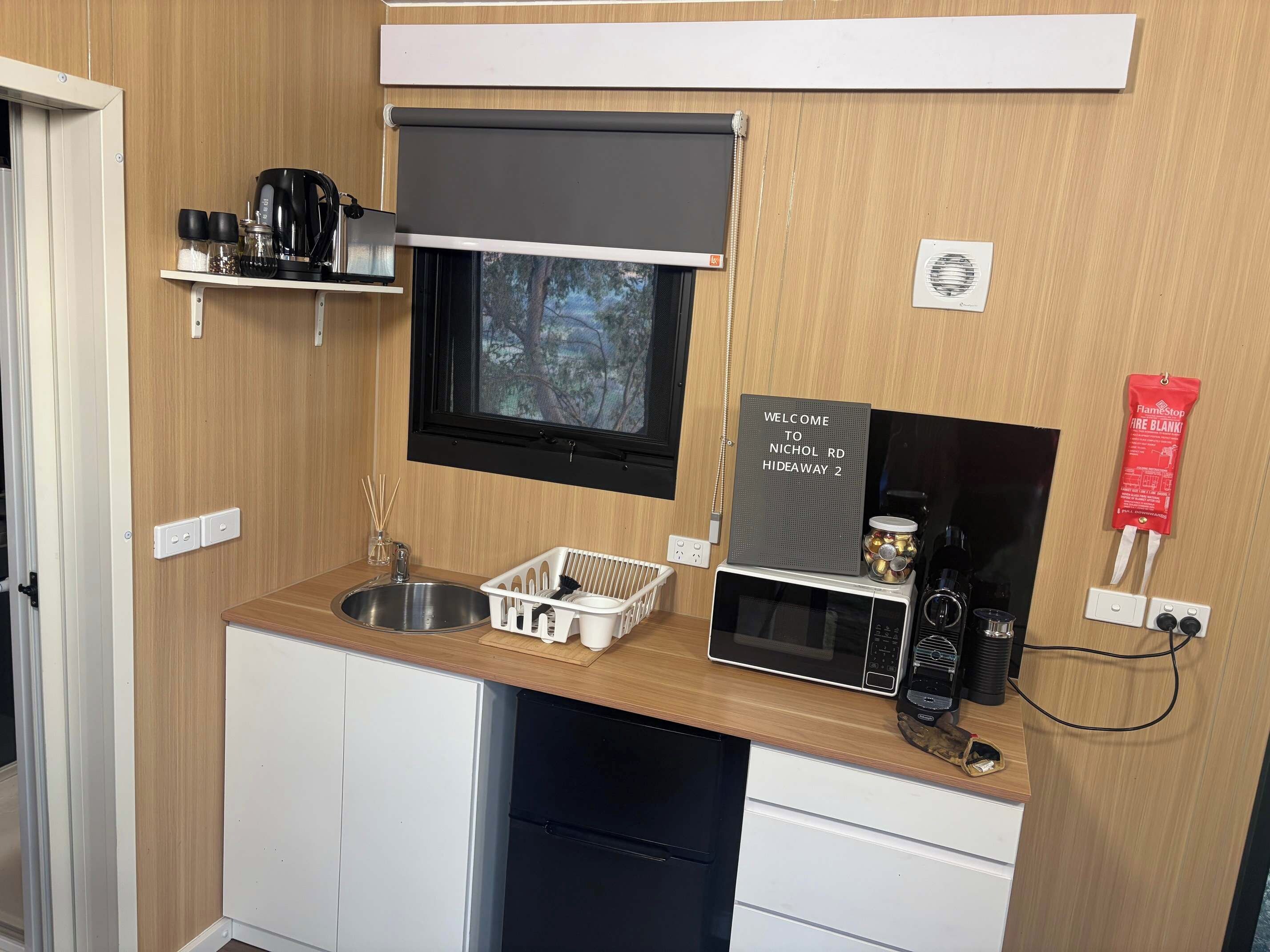 A compact kitchenette with a sink, a microwave, a small refrigerator, a window with a view of trees, and a welcome sign.