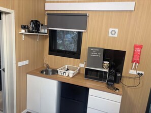Une petite cuisine équipée comprenant un évier, un four à micro-ondes, un petit réfrigérateur, une fenêtre donnant sur les arbres et un panneau d’accueil.