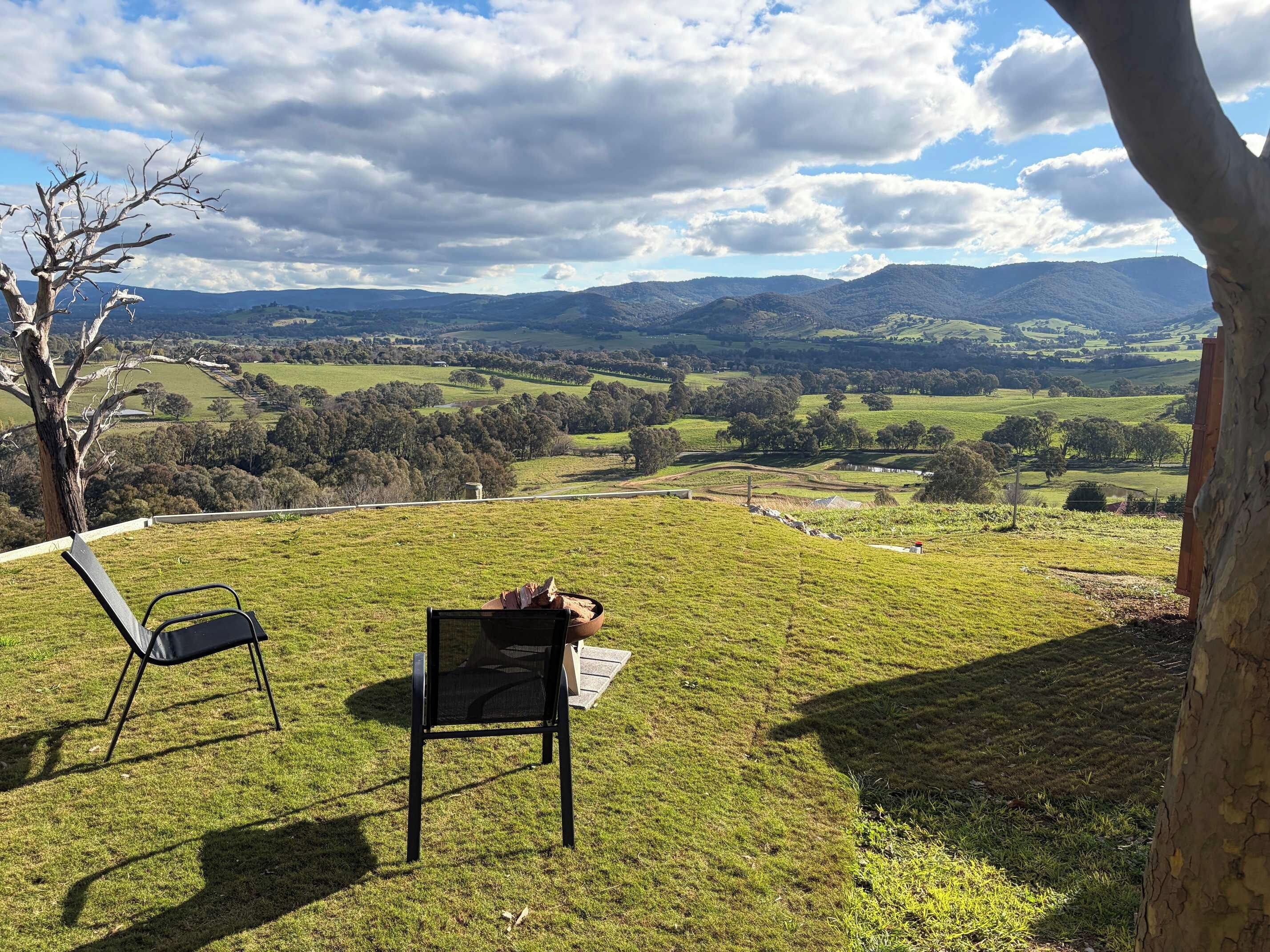 A grassy outdoor area with two chairs and a table, overlooking a valley with hills and trees.