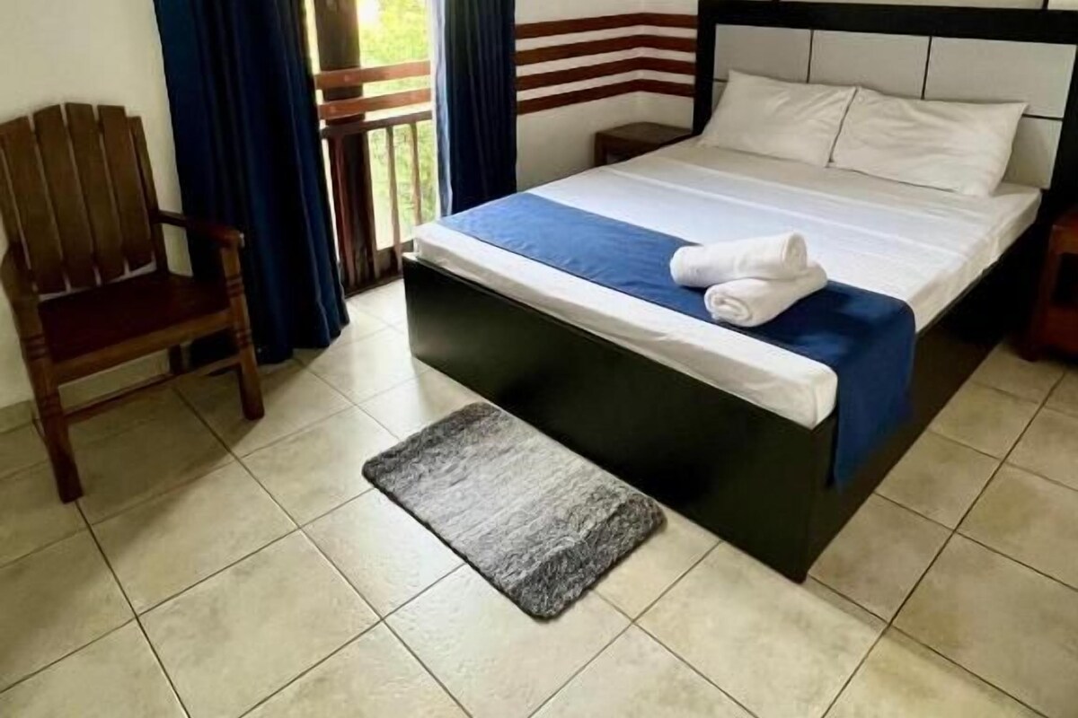 Ein Einzelbett mit Holz Kopfteil, einer blauen Bettdecke und einem weißen Kopfkissen. Außerdem steht ein Holzstuhl und ein kleiner Teppich auf dem Boden.