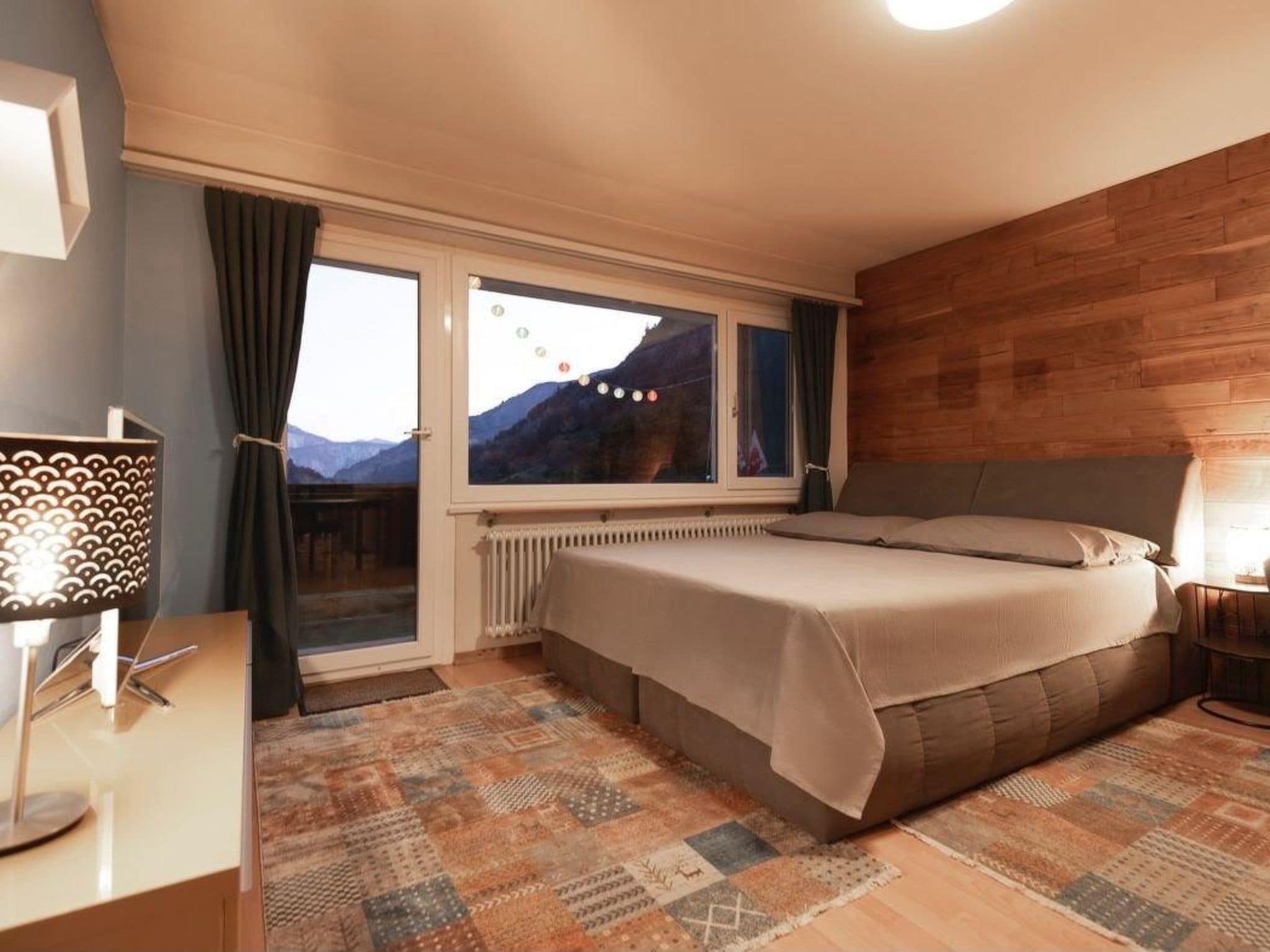 A bedroom with a bed, a window with curtains, a radiator, and a patterned rug.