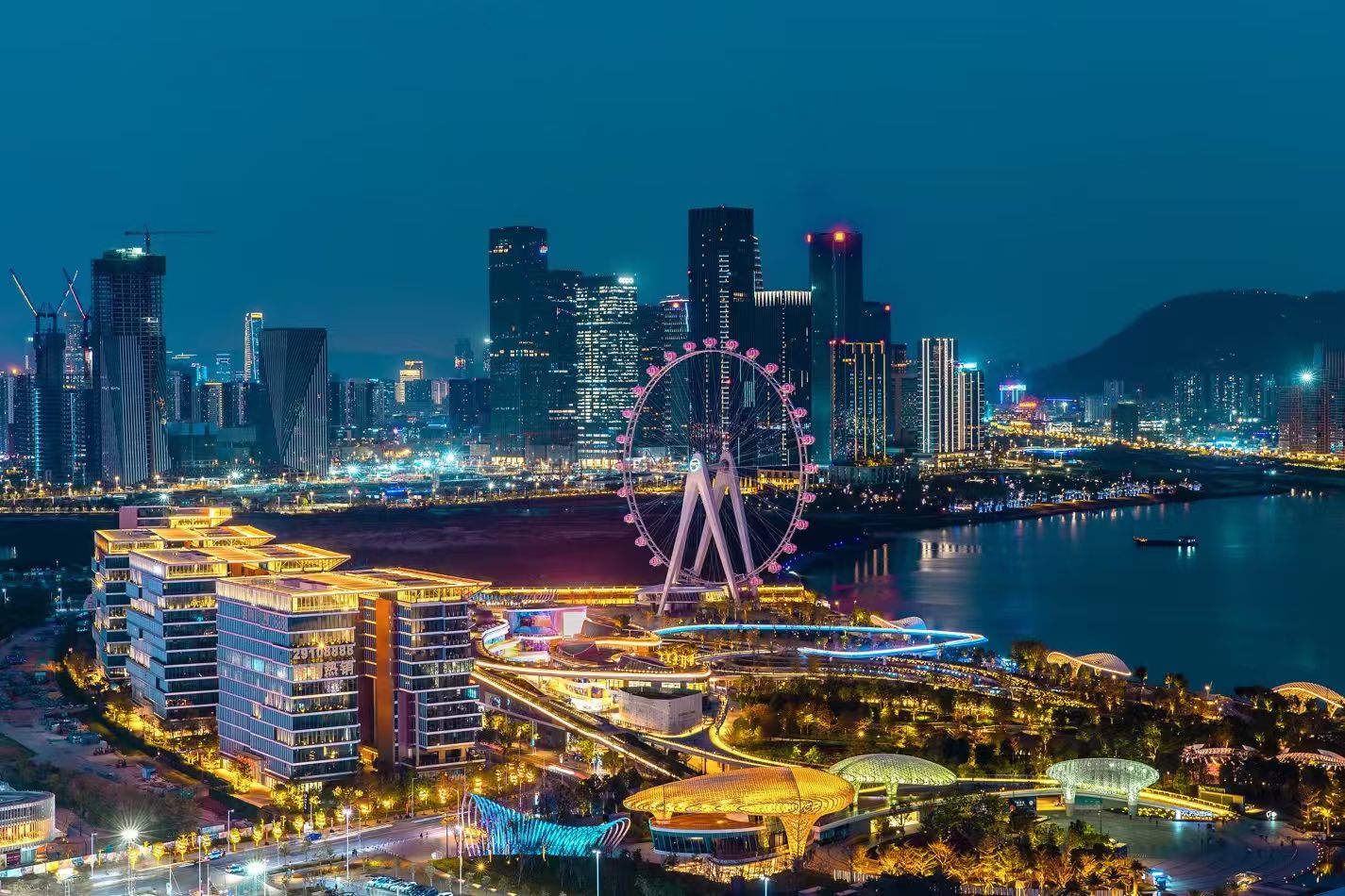 A cityscape at night with a prominent Ferris wheel, high-rise buildings, and a waterfront promenade.