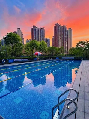 Una piscina con agua cristalina y divisiones para carriles, rodeada de árboles y edificios altos al fondo.