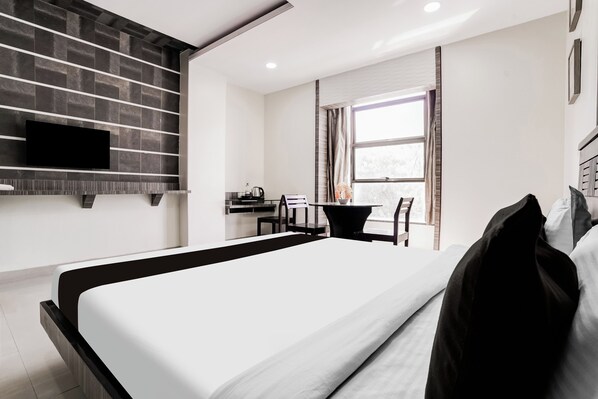 A modern hotel room with a large bed, a flat-screen TV mounted on the wall, a small desk with a chair, and a window with curtains.
