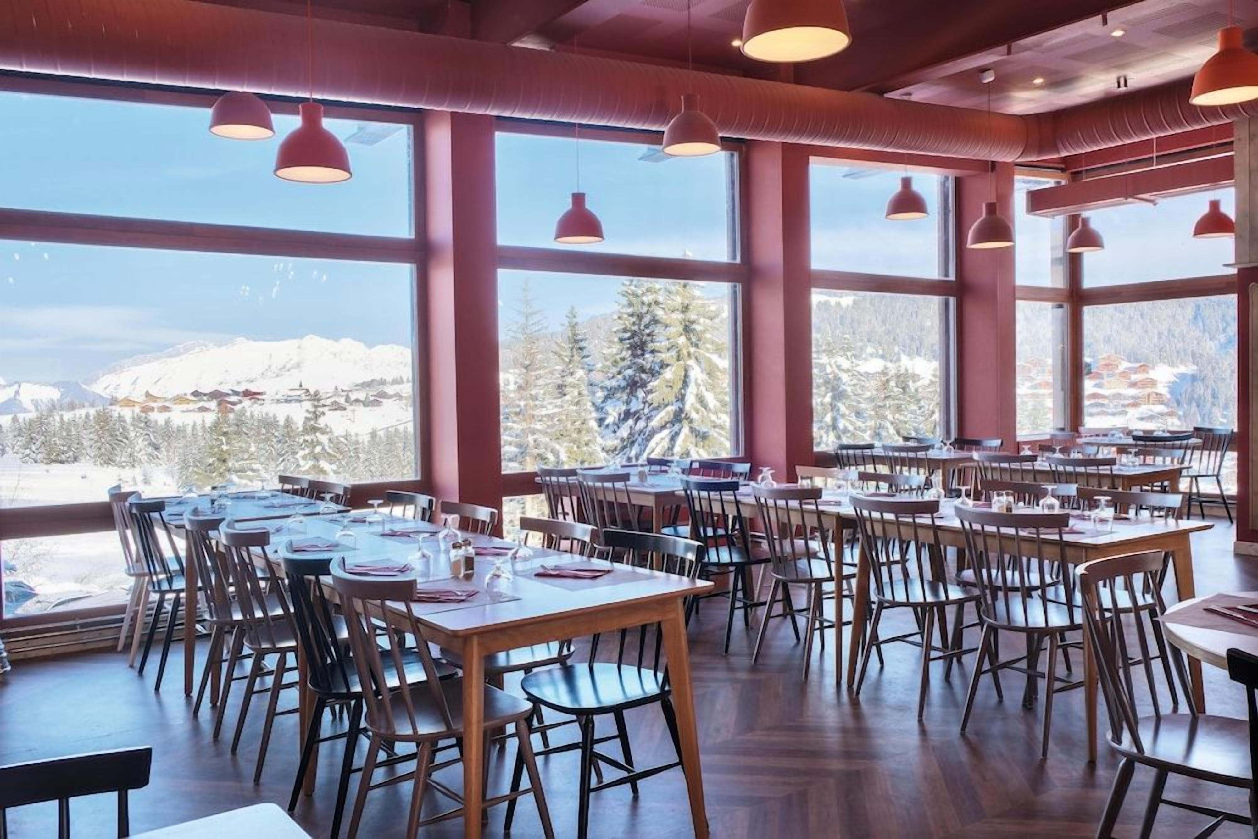 Een restaurant met meerdere tafels en stoelen, grote ramen met uitzicht op een berg en hanglampen.