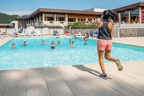 Ett poolområde med folk som badar och solar, en byggnad med en träfasad och en klar himmel.