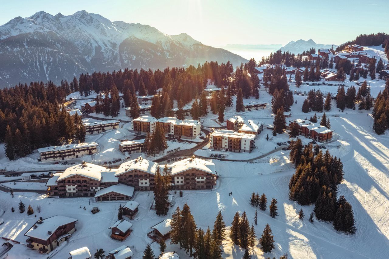 Een bergresort bedekt met sneeuw, met meerdere gebouwen, skipistes en dichte bossen.