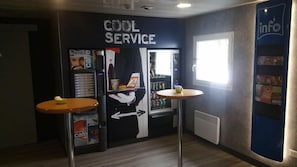 A cafe area with a 'COOL SERVICE' sign, vending machines, and a window providing natural light. - Ibis Budget Flers Grands Champs (Flers)