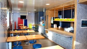 A modern interior with wooden tables and blue chairs, a kitchen area with yellow containers, and a grey wall with a mounted television. - Ibis Budget Flers Grands Champs (Flers)