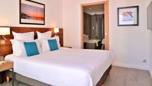 Habitación de hotel con una cama grande, cabecera de madera, mesita de noche, un cuadro con una imagen de un atardecer, y un baño con ducha, además de un cuadro en la pared.